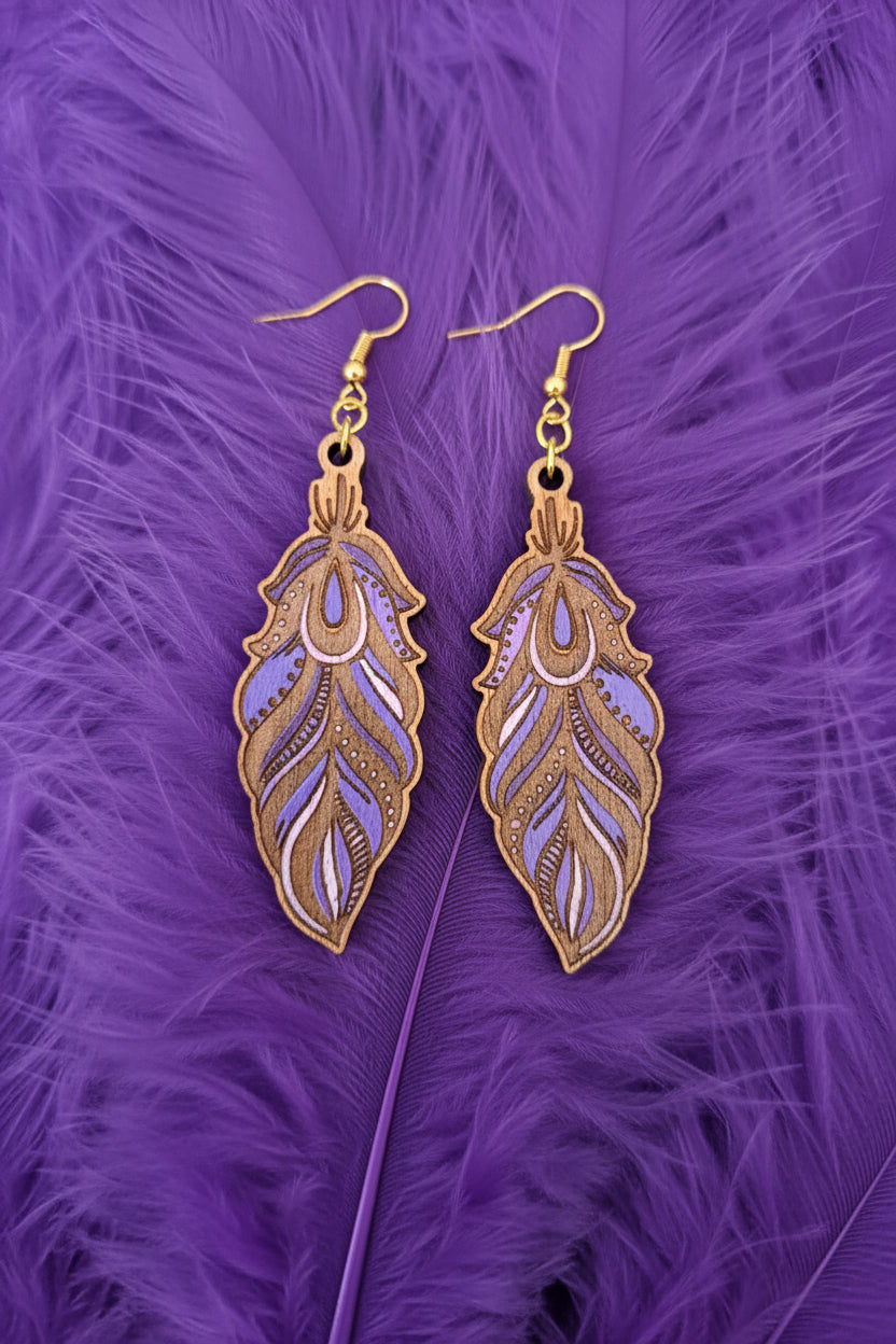A pair of feather-shaped drop earrings with purple and lavender inlay details and gold-tone fishhook wires, displayed on a dark textured surface.