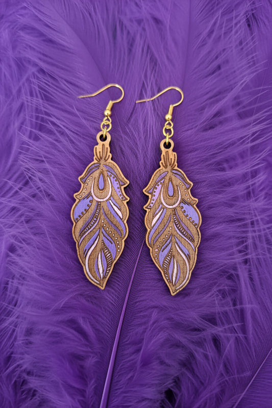 A pair of feather-shaped drop earrings with purple and lavender inlay details and gold-tone fishhook wires, displayed on a dark textured surface.
