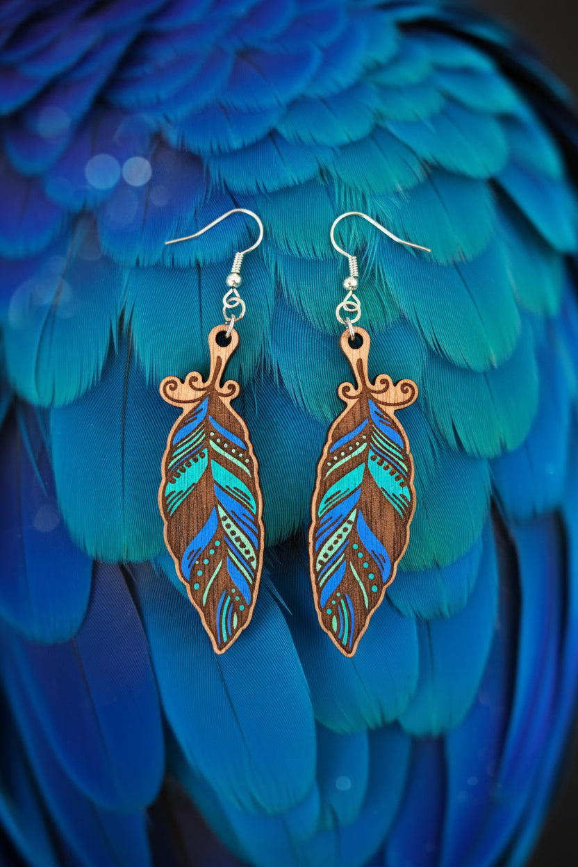 A pair of feather-shaped drop earrings with silver fishhook wires, featuring a brown wood base decorated with turquoise, blue, and black painted patterns, displayed on a dark textured surface.