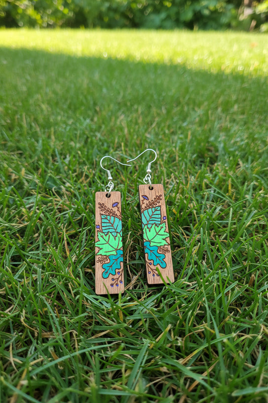 A pair of rectangular wooden drop earrings with hand-painted green and teal leaf designs and silver fishhook ear wires, displayed on a dark textured surface.