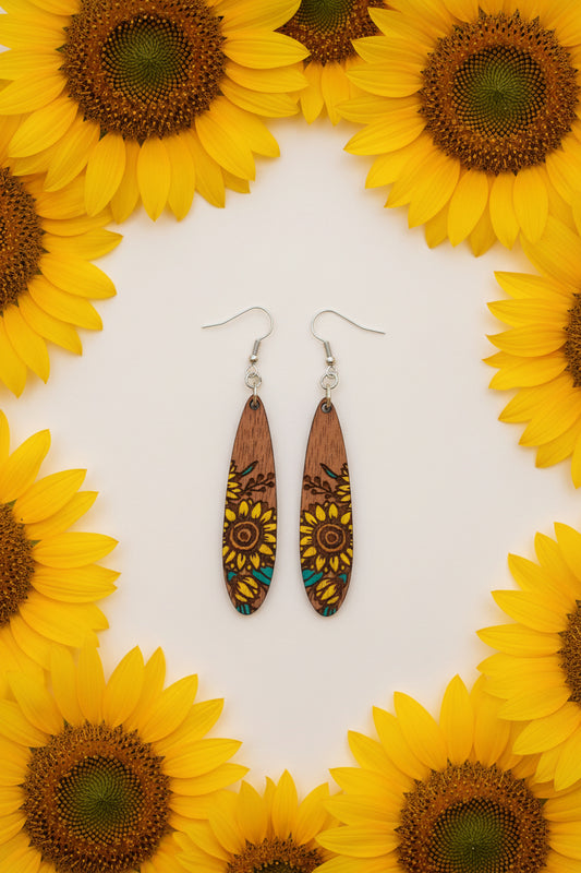 A pair of wooden dangle earrings with engraved sunflower designs, featuring yellow, brown, and teal details, hanging from silver fishhook wires on a dark textured surface.