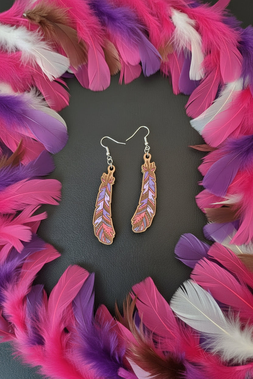 A pair of dangling feather earrings with silver fishhook wires. The feather charms are brown leather or wood with colorful hand-painted patterns in pink, purple, and light lavender.